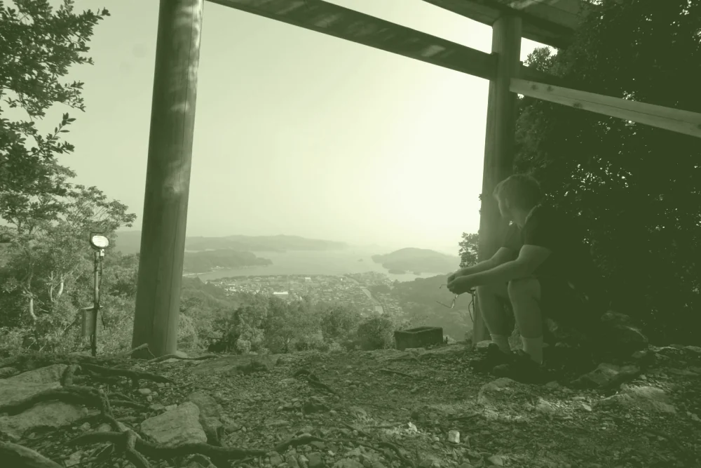 Aodren Le Pesquer assis sous un torii, en surplomb d'une baie dans la brume.